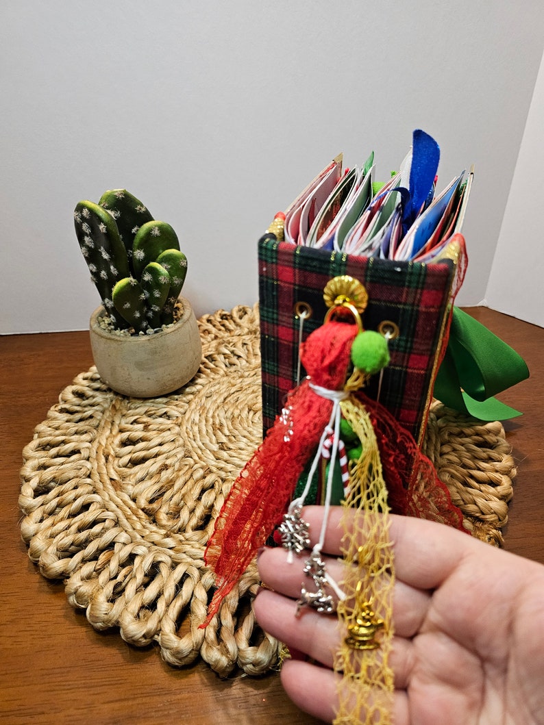 May include: A red and green plaid fabric covered journal with a red ribbon, white lace, and green and gold embellishments. The journal is decorated with a small, white, and red pom-pom.