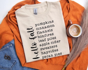 May include: A beige t-shirt with the words "hello fall" in a script font and a list of fall-themed items in a black sans-serif font. The shirt is on an orange sweater, with a cup of coffee and a pair of jeans.