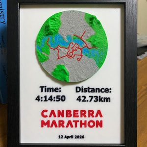 May include: Framed artwork with a 3D map of a city, showing green and grey terrain, blue water, and red pathways. The text "CANBERRA MARATHON" is displayed, along with the time 4:14:50 and distance 42.73km. The date "12 April 2026" is also included.