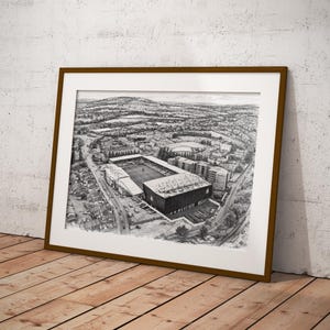 May include: A framed black and white print of a stadium, viewed from above. The artwork is set against a white mat and framed in a brown wooden frame. The print is leaning against a white wall and wooden floor.