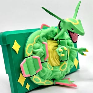 Może przedstawiać: Wydrukowana w 3D zielona figurka Rayquazy z żółtymi i różowymi akcentami. Figurka jest zamontowana na zielonej prostokątnej podstawie z żółtymi dekoracjami w kształcie gwiazd. Rayquaza ma szczegółowy design z otwartą paszczą i wyciągniętymi ramionami.