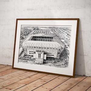 Puede incluir: Un dibujo arquitectónico en blanco y negro enmarcado de un estadio, apoyado contra una pared blanca y un suelo de madera. El dibujo detallado muestra la estructura del estadio, los edificios circundantes y los estacionamientos. El marco es marrón.