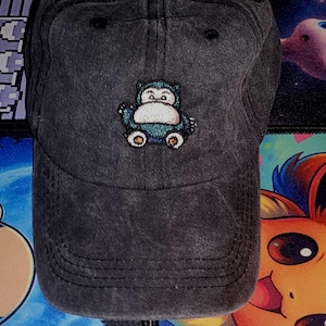 May include: A dark gray baseball cap featuring an embroidered Snorlax design. The cap has a curved brim and an adjustable strap. The Snorlax design is in shades of blue, white, and brown.