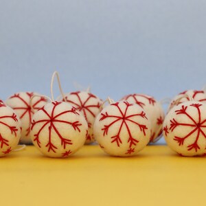 May include: A set of ten white felt Christmas baubles with red snowflake designs. The baubles are arranged in a row on a yellow surface.