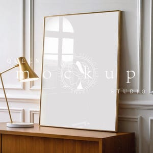 Pode incluir: Maquete de impressão de arte retangular com moldura dourada e interior branco. A moldura está sobre um armário de madeira, ao lado de uma luminária de mesa dourada. As palavras "QUEEN MOCKUP STUDIO" estão impressas.