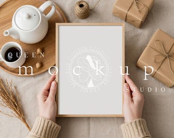 May include: A wooden picture frame held by hands, with the text "QUEEN MOCKUP STUDIO" and a logo. The frame is surrounded by a teapot, cup, gifts, and decorative items, creating a cozy, neutral-toned aesthetic.