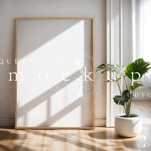 May include: A large, empty, wooden picture frame with a white mat is leaning against a white wall. The frame is in a room with a white floor and a potted plant with large green leaves.