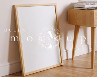 May include: A large, blank white poster in a light wood frame, leaning against a wall. The frame is rectangular and the poster is centered. A wooden side table with books is to the right. The floor is wood.