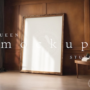 May include: A large, ornate gold picture frame with a white mat and a blank canvas inside. The frame is standing on a wooden floor in front of a wood-paneled wall. A small table with a stack of books and a potted plant is in the foreground.