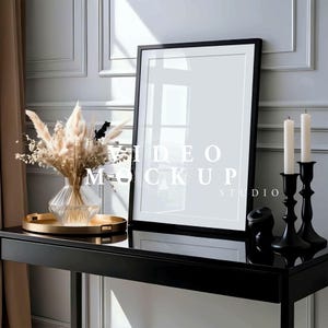 Könnte beinhalten: Ein schwarzer Bilderrahmen mit weißem Passepartout steht auf einem schwarzen Tisch. Eine klare Vase mit Trockenblumen steht auf einem goldenen Tablett. Zwei schwarze Kerzenhalter mit weißen Kerzen stehen auf dem Tisch.
