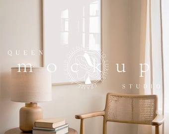 May include: A beige lamp with a linen shade sits on a wooden table next to a stack of books. A wooden chair with a woven seat sits in the corner of the room. A large framed picture hangs on the wall behind the chair. The text "QUEEN MOCKUP STUDIO" is visible in the image.
