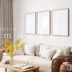 May include: Three square frames with a white background and a light wood frame hang on a white wall above a white couch with white pillows. The couch is in front of a window with a sheer beige curtain. A vase with yellow flowers sits on a wooden coffee table in front of the couch.