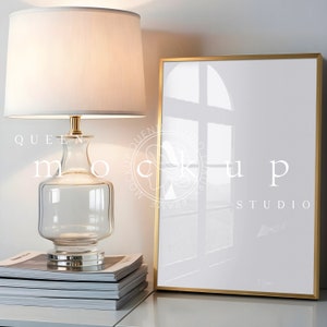 May include: A gold-framed picture frame with a white mat and a white background. The frame is sitting on a white surface with a stack of magazines and a glass lamp with a white lampshade.