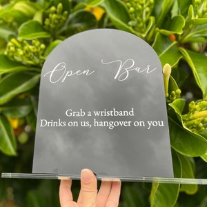 May include: A grey acrylic sign with a semi-circular top, featuring the words "Open Bar" in white script. The sign also reads "Grab a wristband, Drinks on us, hangover on you." It is supported by a clear acrylic base, ideal for events.