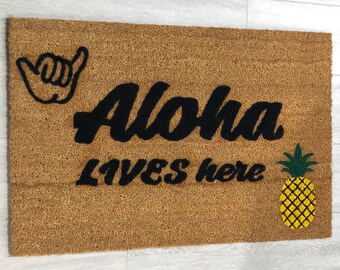 Op de afbeelding: Een bruine kokosvezel deurmat met de tekst "Aloha Lives Here" in zwarte cursieve letters. Een zwart-wit handgebaar en een gele ananas met groene bladeren zijn ook op de mat.