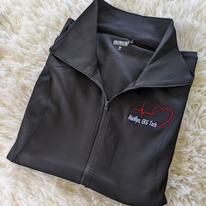 May include: Black zippered jacket with a red heart and EKG graphic embroidered on the front. The text "Madilyn, EKG Tech" is embroidered below the graphic.