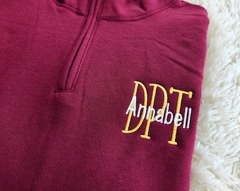 May include: Burgundy quarter-zip sweatshirt with a gold embroidered monogram "DPT" and the name "Annabell" in white.