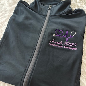 May include: Black zippered jacket with a purple embroidered heart, stethoscope, and EKG line. The text "Krupali, RDMS Cardiovascular Sonographer" is also embroidered on the jacket.
