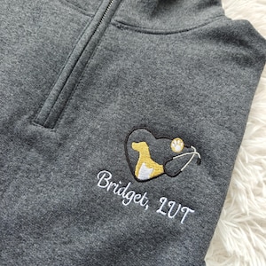 Peut inclure: Un sweat-shirt gris à fermeture éclair 1/4 avec un cœur brodé contenant une silhouette de chien et de chat, un stéthoscope et le texte "Bridget, LVT".