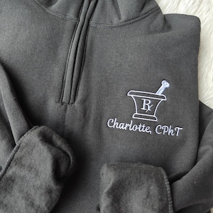 Peut inclure: Un sweat-shirt gris foncé à fermeture éclair 1/4 avec un mortier et un pilon brodés en blanc avec un "R" à l'intérieur et le texte "Charlotte, CPhT".