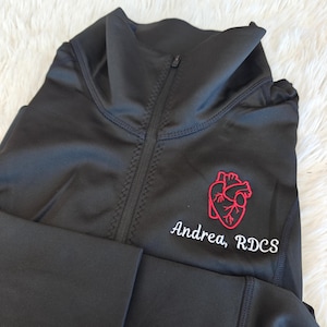 May include: Black long-sleeve zip-up shirt with a red embroidered heart and the text "Andrea, RDCS" in white.