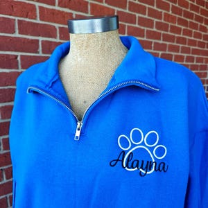 May include: A royal blue sweatshirt with a quarter-zip collar. The sweatshirt features a white paw print design with the name "Alayna" in a script font. The zipper is silver.