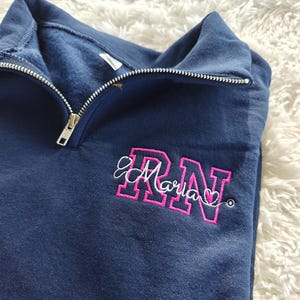 May include: A navy blue 1/4 zip sweatshirt with a pink embroidered design that says "RN" with the name "Maria" written in cursive underneath.