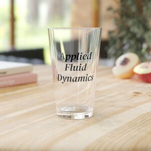Pode incluir: Um copo de cerveja transparente com as palavras "Applied Fluid Dynamics" impressas em preto. O copo está sobre uma superfície de madeira clara, com um fundo desfocado.