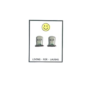 May include: A pair of small, gray tombstone earrings with the text "R.I.P." on each. The earrings are on a white background with a yellow smiley face above them. The text "LIVING FOR LAUGHS" is below the earrings.