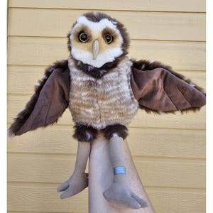 May include: A plush owl toy with brown and white fur, large dark eyes, and a light-colored beak. The toy features outstretched wings, long tan fabric legs, and a small blue band around one leg, showcasing a detailed and realistic bird design.
