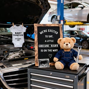 May include: A baby announcement in a garage setting. A white onesie with "Baby Thompson September 2020" hangs above a toolbox. A teddy bear in denim overalls holds a wrench next to a sign that reads "We're excited to say, a little grease baby is on the way!"