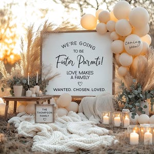 May include: A wooden sign with the text "We're going to be Foster Parents! Love makes a family. Wanted. Chosen. Loved."  The sign is surrounded by pampas grass, beige balloons, and candles. A smaller sign reads "The Thompson Family".