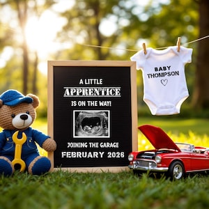Op de afbeelding: Een zwart schoolbord met de tekst "A Little Apprentice is on the way! Joining the Garage February 2026". Een teddybeer met een blauwe monteursoutfit en een moersleutel zit naast het schoolbord. Een witte romper met de tekst "Baby Thompson" hangt aan een waslijn. Een rode speelgoedauto met de motorkap open staat op de voorgrond.