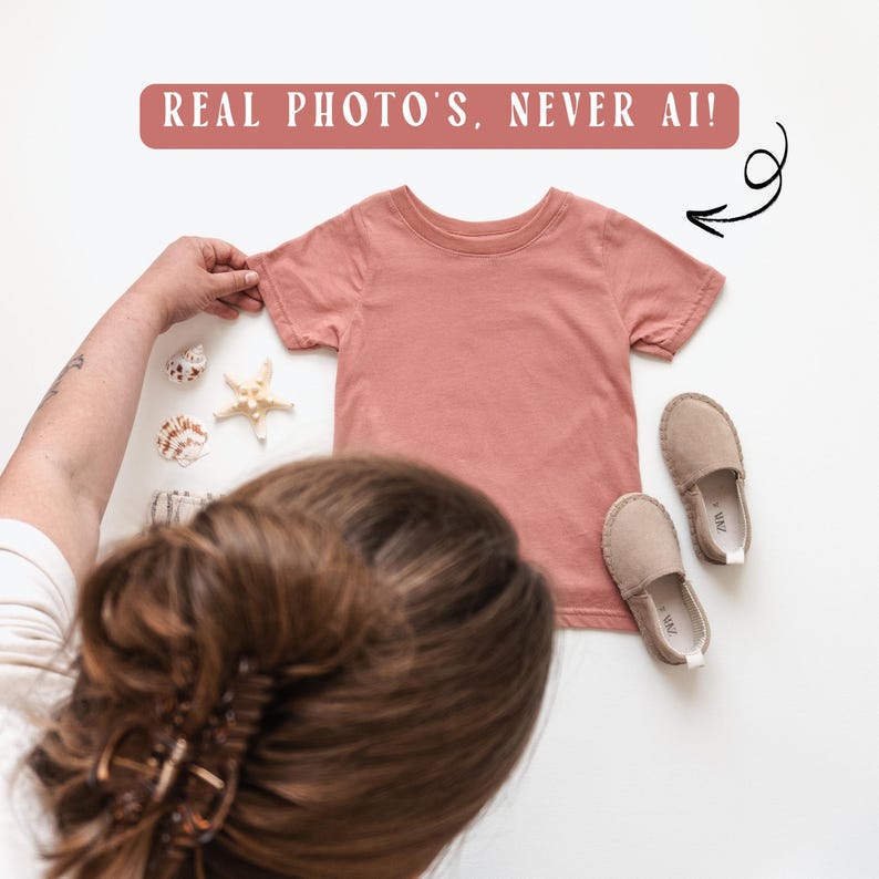 May include: A coral-coloured t-shirt with short sleeves, displayed with a pair of beige shoes and seashells. The text "REAL PHOTO'S, NEVER AI!" is above the shirt. The shirt is likely for children.
