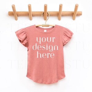 Puede incluir: Una camiseta de color rosa polvoriento con mangas cortas de volantes cuelga de una percha de madera. La camiseta tiene el texto "your design here" impreso en la parte delantera.
