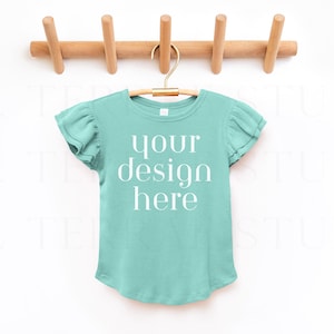 Puede incluir: Una camiseta azul turquesa con mangas de volantes colgada de una percha de madera. La camiseta tiene el texto "your design here" impreso en la parte delantera.