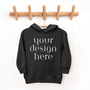 May include: Black hooded sweatshirt hanging on a wooden rack with the text "your design here" printed on the front.