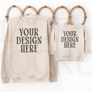 Könnte beinhalten: Zwei beige Sweatshirts, eine Erwachsenen- und eine Kindergröße, hängen an einer hölzernen Wanddekoration. Auf jedem Sweatshirt steht in dunkelgrauer Schrift "YOUR DESIGN HERE". Die Sweatshirts hängen an goldenen Kleiderbügeln.