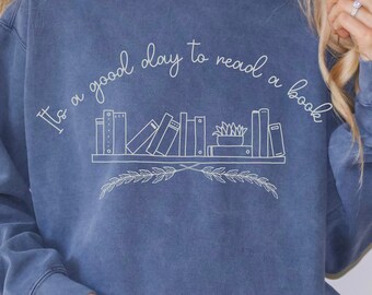 May include: A blue sweatshirt with the text "It's a good day to read a book" and a graphic of a bookshelf with books and a plant.