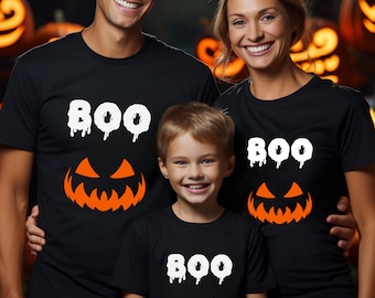 Puede incluir: Una familia de tres personas con camisetas negras con un "BOO" blanco y una cara de calabaza naranja. Las camisetas son una forma divertida y festiva de celebrar Halloween.