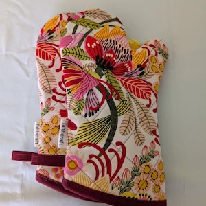 May include: Quilted oven gloves featuring a colourful floral design in red, pink, yellow, and green on a cream backdrop. The gloves have a burgundy trim and a hanging loop. The tag reads "faithcraft".