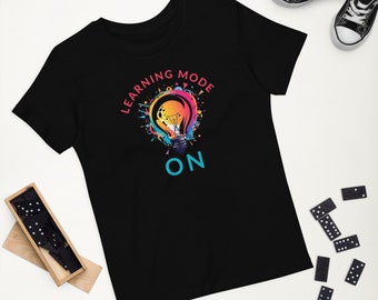 Puede incluir: Una camiseta negra de manga corta con un gráfico de una bombilla colorida con salpicaduras de pintura. El texto "LEARNING MODE" está arqueado sobre la bombilla en rosa, y la palabra "ON" está impresa en azul claro bajo el diseño.