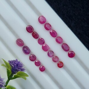 May include: A collection of 15 oval-shaped gemstones in various shades of pink, arranged in rows on a white surface.