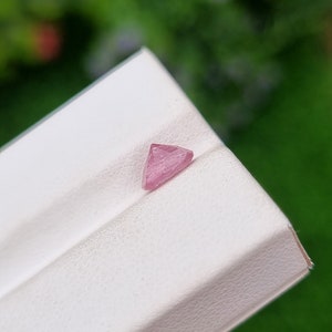 May include: A pink triangular gemstone with facets, displayed on a white surface.
