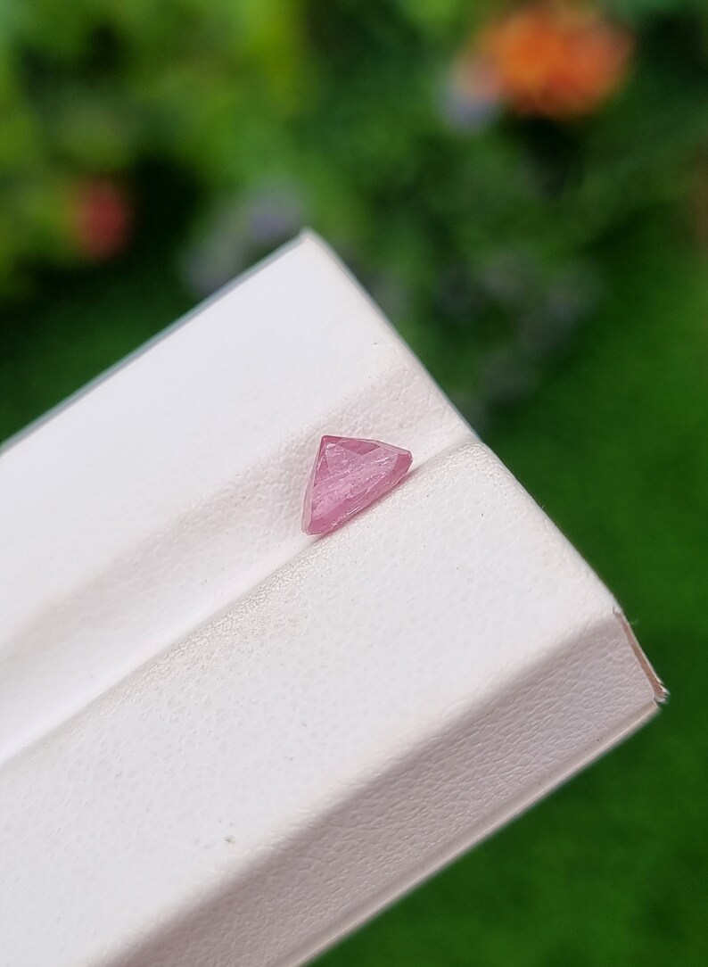 May include: A pink triangular gemstone with facets, displayed on a white surface.