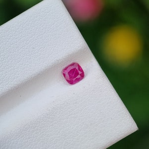 Puede incluir: Una gema rosa vibrante, probablemente un rubí, se muestra sobre una superficie blanca texturizada. La gema tiene forma cuadrada y un acabado pulido.