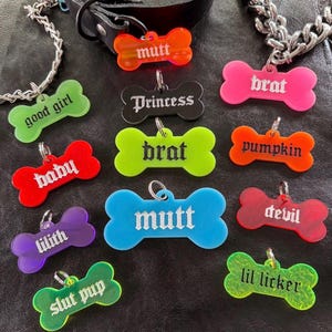 May include: A collection of 12 colorful bone-shaped dog tags with various names and phrases printed on them, including "mutt", "good girl", "princess", "brat", "baby", "lilith", "pumpkin", "devil", "slut pup", and "lil licker".