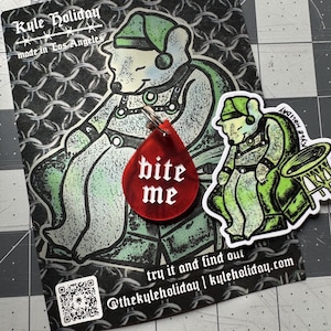 May include: A red, teardrop-shaped acrylic pendant featuring the white text "bite me" in a gothic font. The pendant is displayed on a black and white backing card with a graphic illustration of a bear and the text "Kyle Holiday".