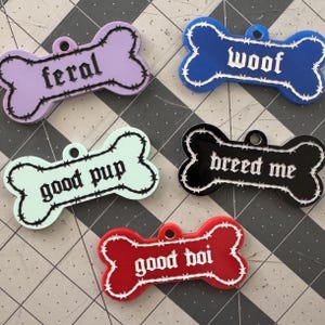 May include: Five bone-shaped pet tags in various colors: purple with "feral", blue with "woof", mint green with "good pup", black with "breed me", and red with "good boi". Each tag has a white outline and barbed wire design.