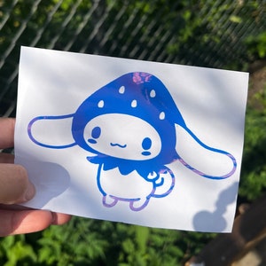 May include: A blue and purple iridescent vinyl sticker featuring a cartoon character with a strawberry on its head. The character has large ears and a small, smiling mouth.